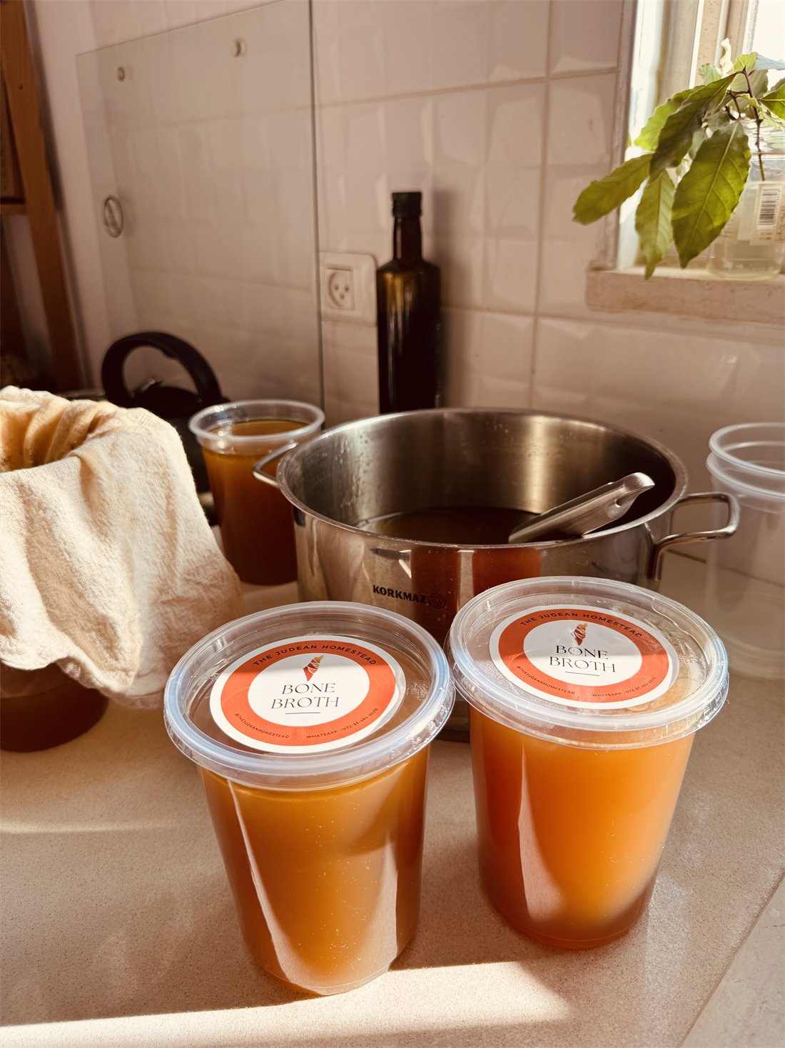 Freshly labeled jars of The Judean Homestead bone broth on the counter beside the simmering pot
